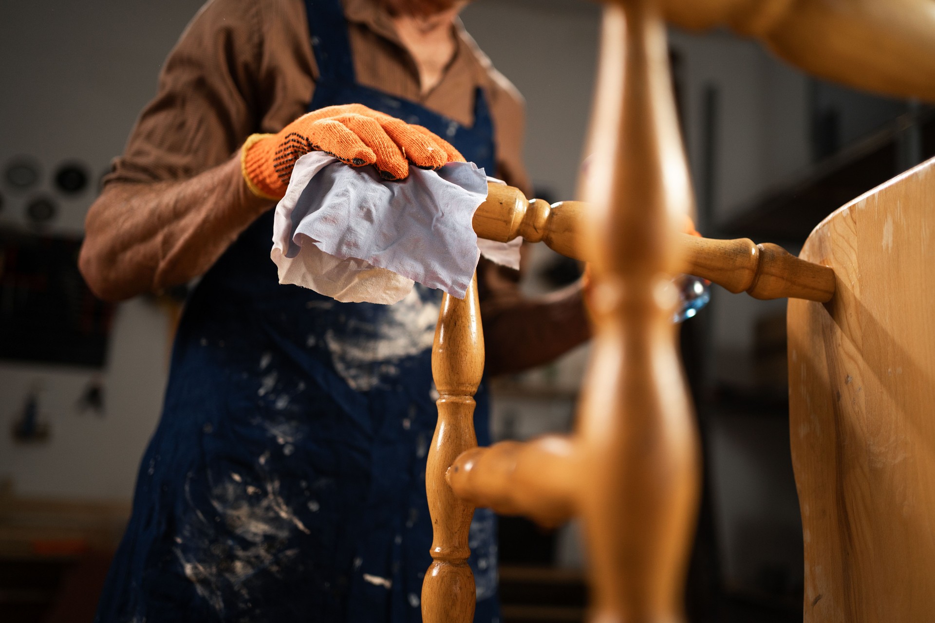 An elderly man treats a wooden chair with a degreaser in his workshop. He is wearing a brown shirt, blue apron and orange gloves, carefully cleaning the chair. concept restoration of old furniture